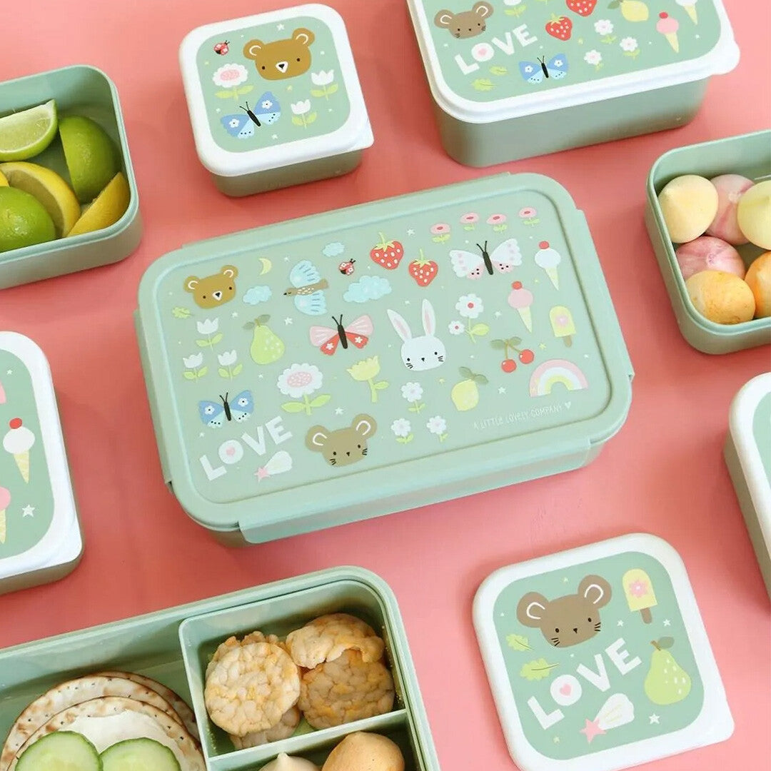 Een collectie van a little lovely company broodtrommels en snackdoosjes voor kinderen, gevuld met eten, op een roze achtergrond.
