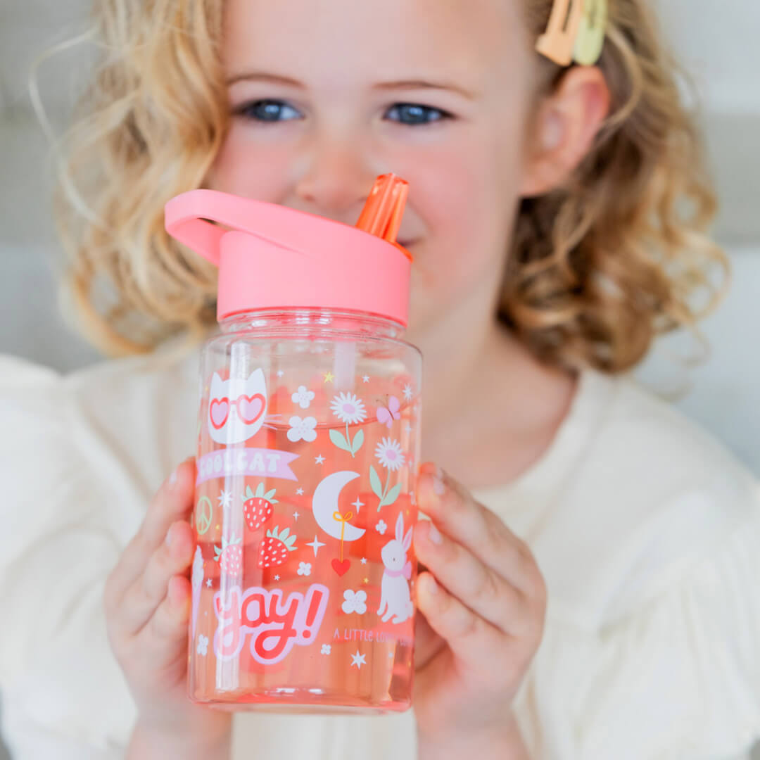 Een kind houdt de a little lovely company kinderdrinkfles met roze deksel en rietje vast, gevuld met roze vloeistof en versierd met 'COOL CAT' en een konijn.