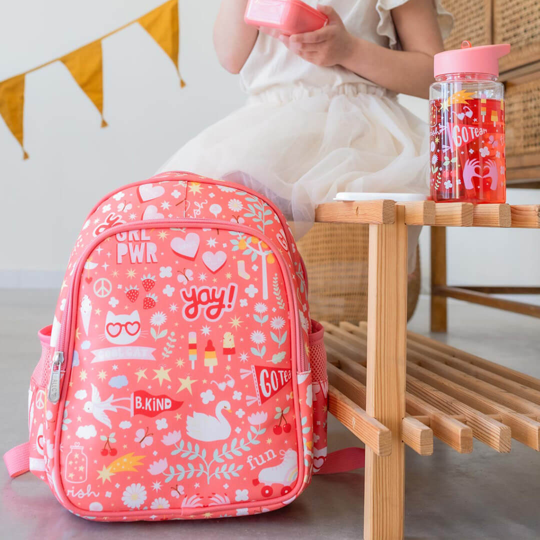 Een roze rugzak met speelse figuren staat naast een houten bankje, met de a little lovely company kinderdrinkfles ernaast.