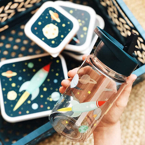 Een hand houdt de a little lovely company drinkfles met rietje met ruimte-thema vast, met bijpassende lunchboxen op de achtergrond.