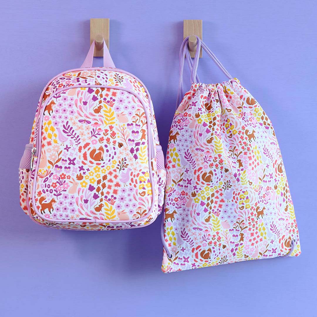 a little lovely company kinderrugzak en gymtas met bloemen- en dierenprint hangen aan een paarse muur.