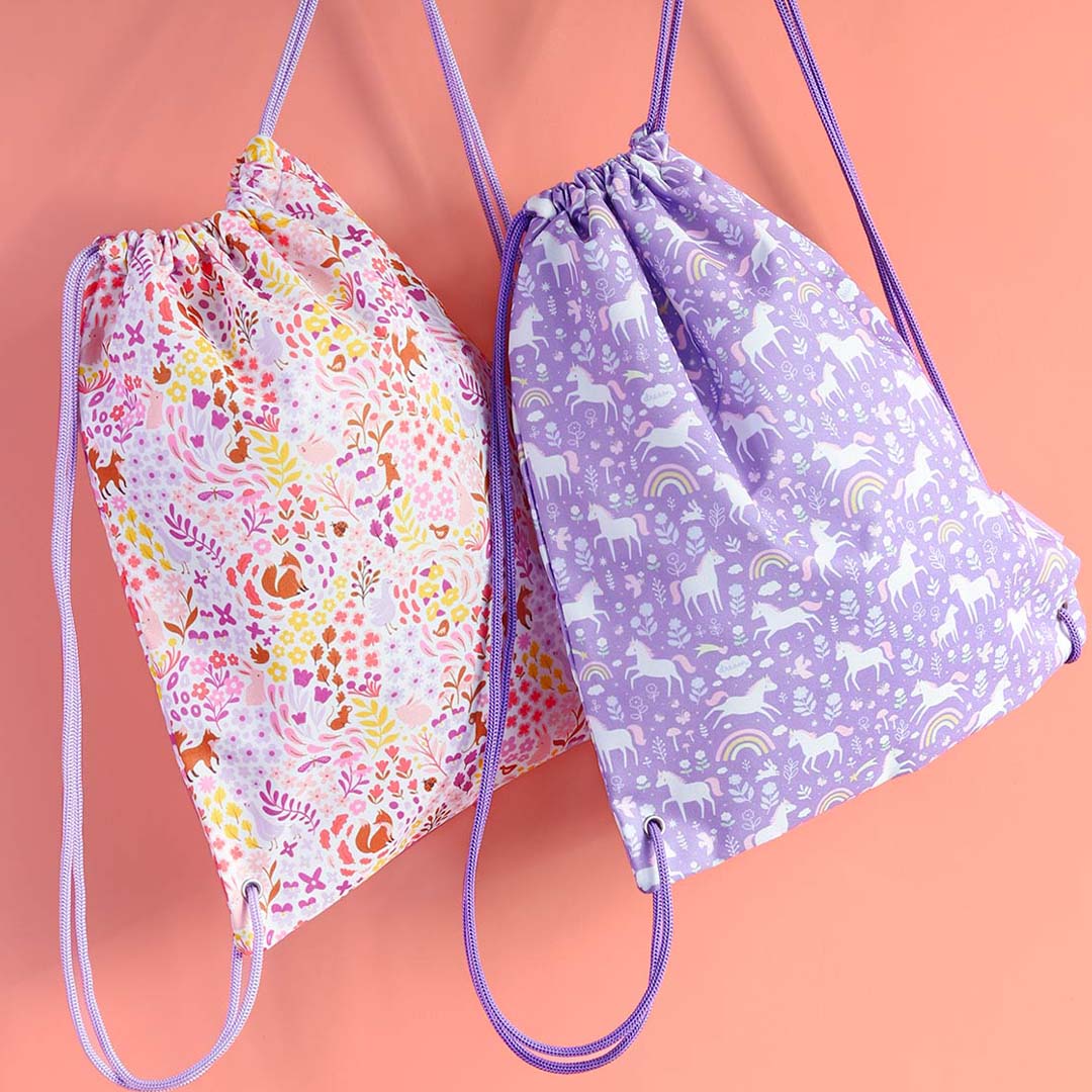 Twee gymtassen hangen naast elkaar, één paars met eenhoorns en één roze met bloemen en dieren.