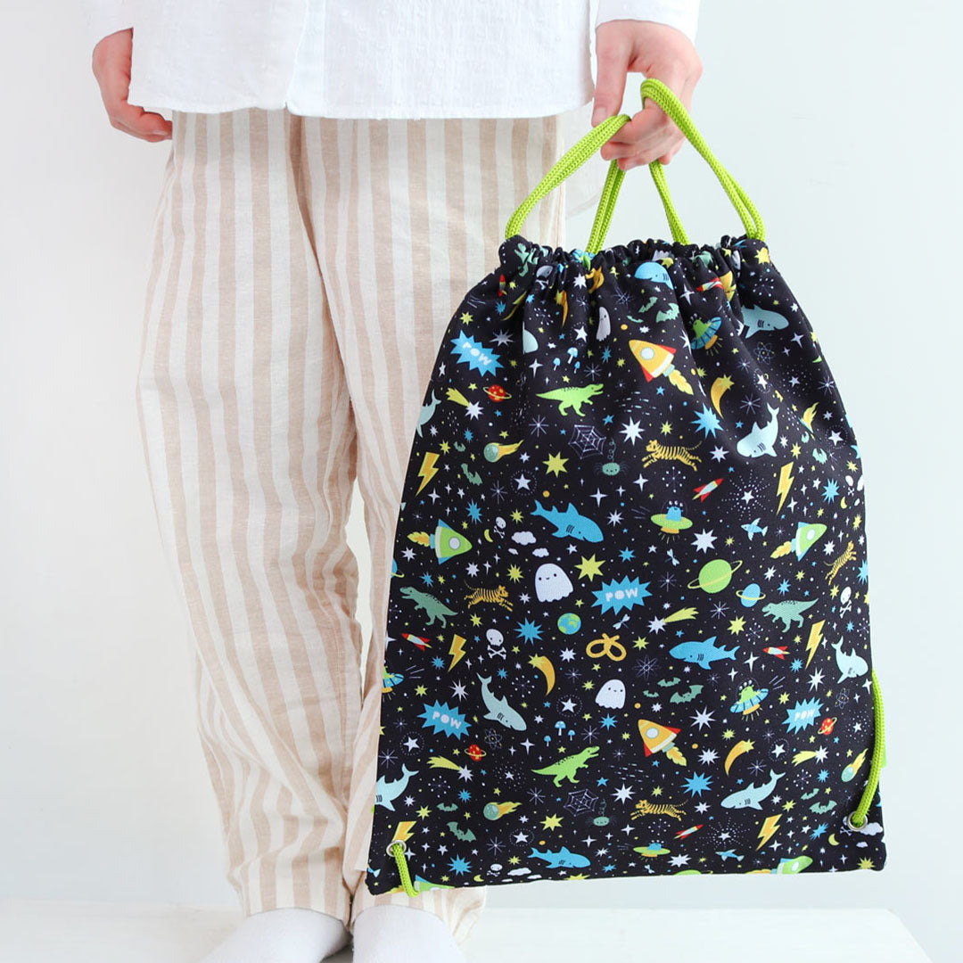 Een persoon in pyjama houdt de a little lovely company zwarte kindergymtas met galaxy- en dierenpatroon vast.