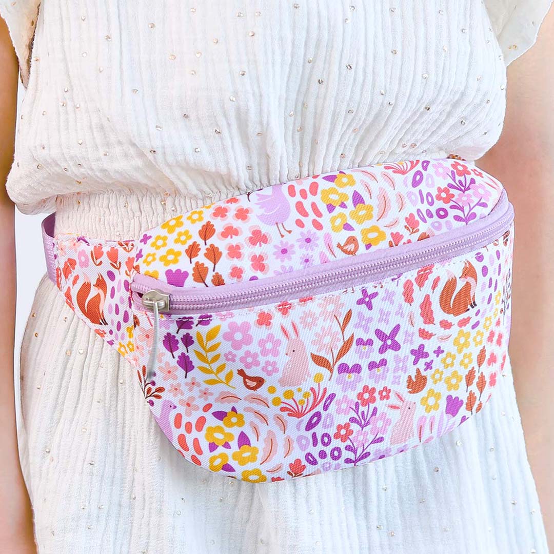 Een a little lovely company kinder heuptas met bloemen- en dierenprint wordt gedragen om de taille van een persoon in een witte jurk.