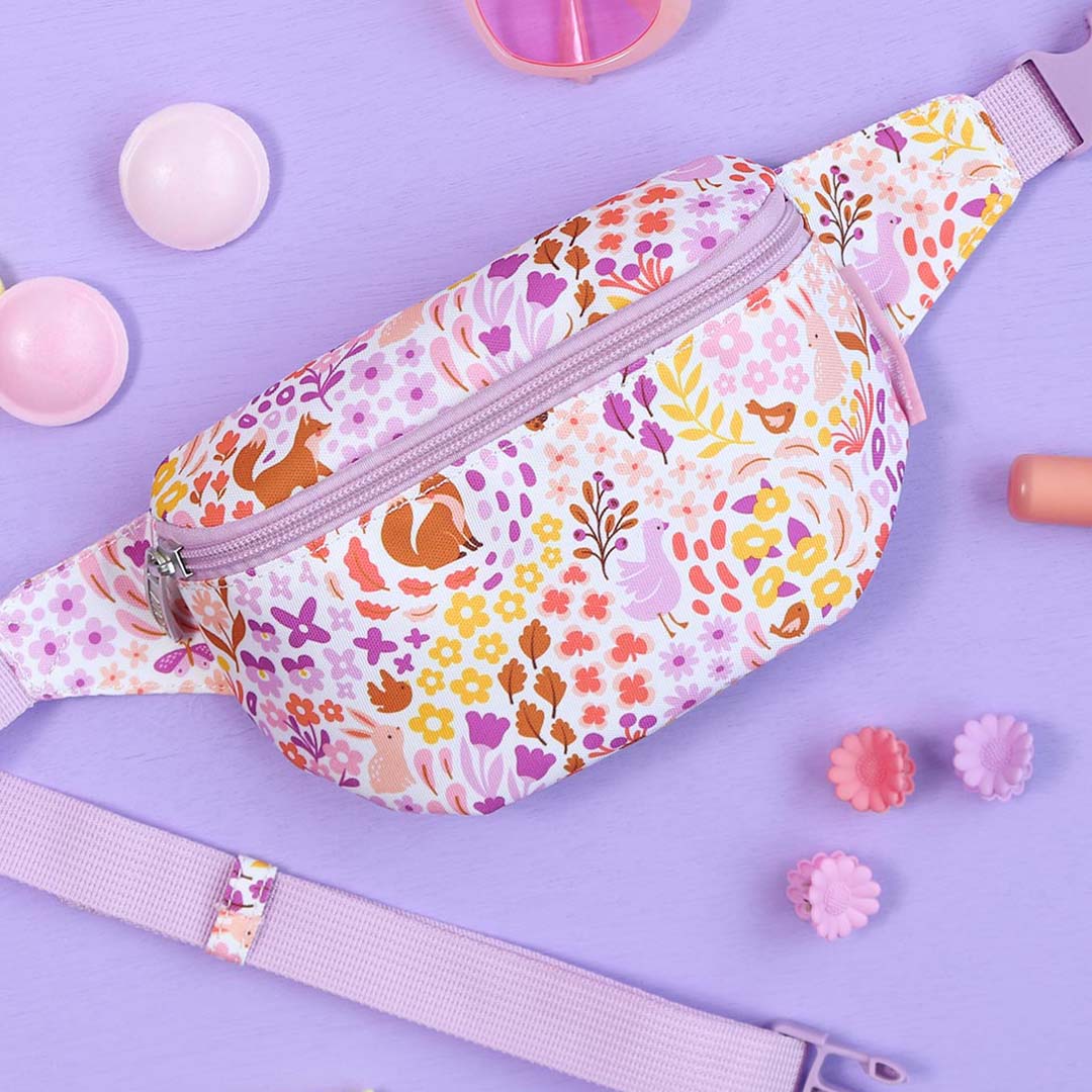 Een a little lovely company kinder heuptas met bloemen- en dierenpatroon ligt op een paarse ondergrond met roze speelgoedaccessoires.