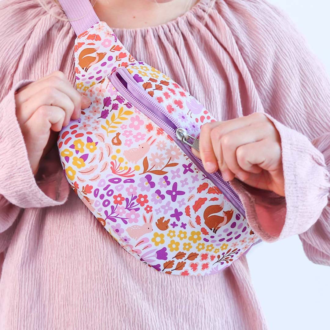 Een a little lovely company kinder heuptas met bloemen- en dierenprint wordt door kinderhanden geopend terwijl deze over de borst wordt gedragen.