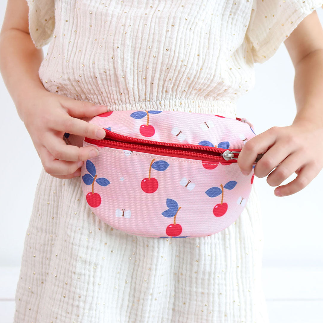 Een kind draagt de roze a little lovely company kinderheuptas met kersenprint om de taille en opent de rits.