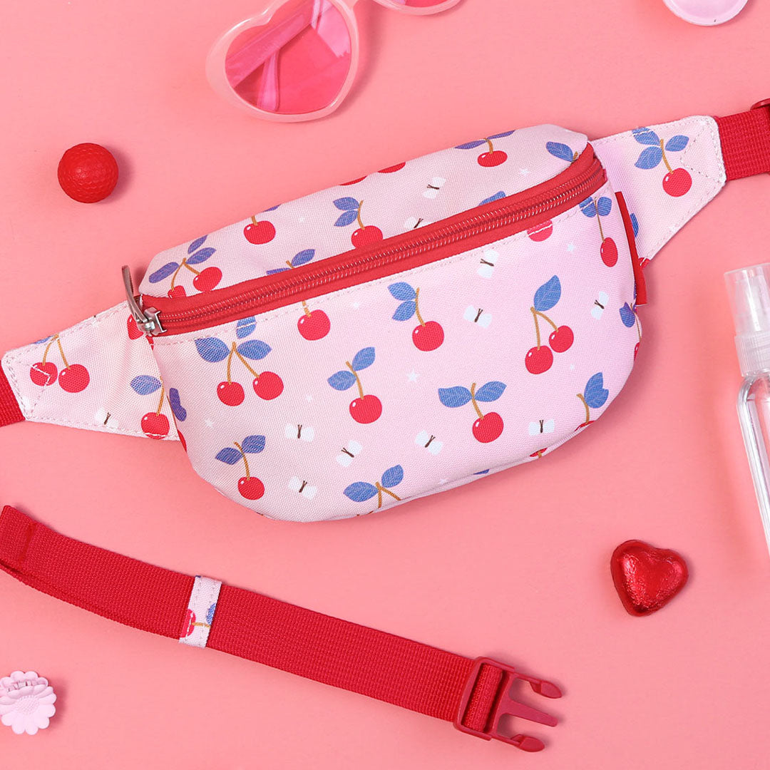 De roze a little lovely company kinderheuptas met kersenprint ligt op een roze ondergrond tussen roze accessoires.