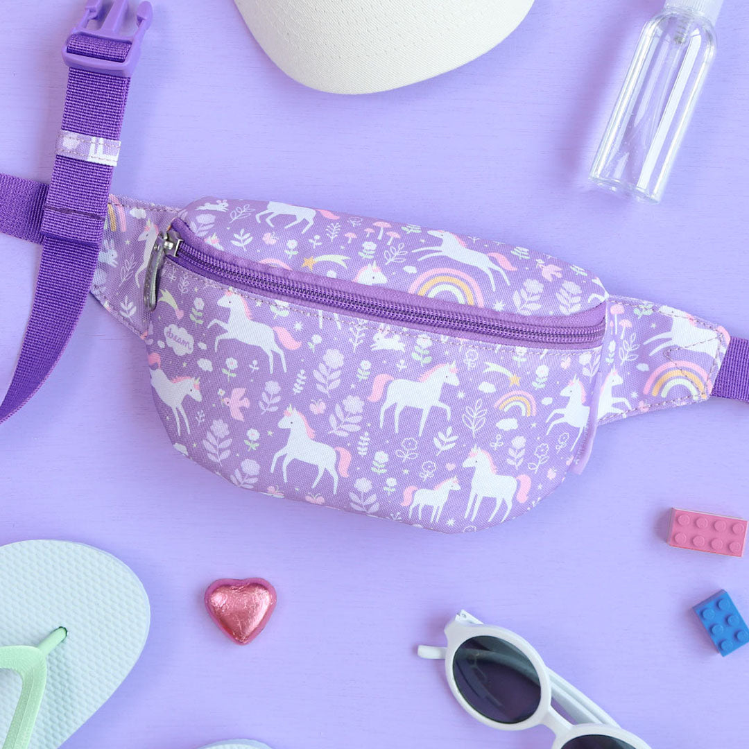 De paarse kinder heuptas met eenhoorn- en regenboogpatroon ligt op een paarse ondergrond tussen zomerse accessoires.