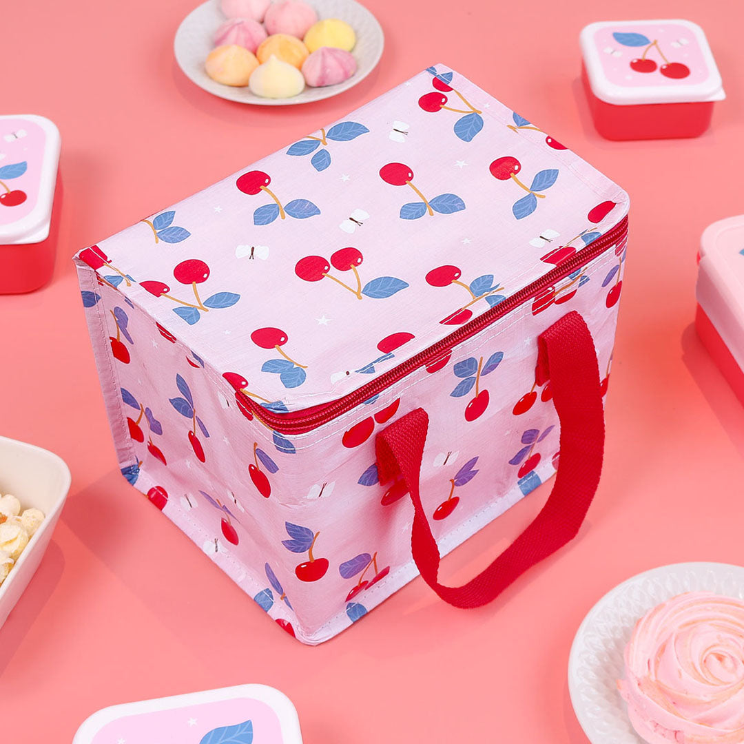 De a little lovely company roze koeltas met kersenpatroon staat op een roze ondergrond tussen snacks.