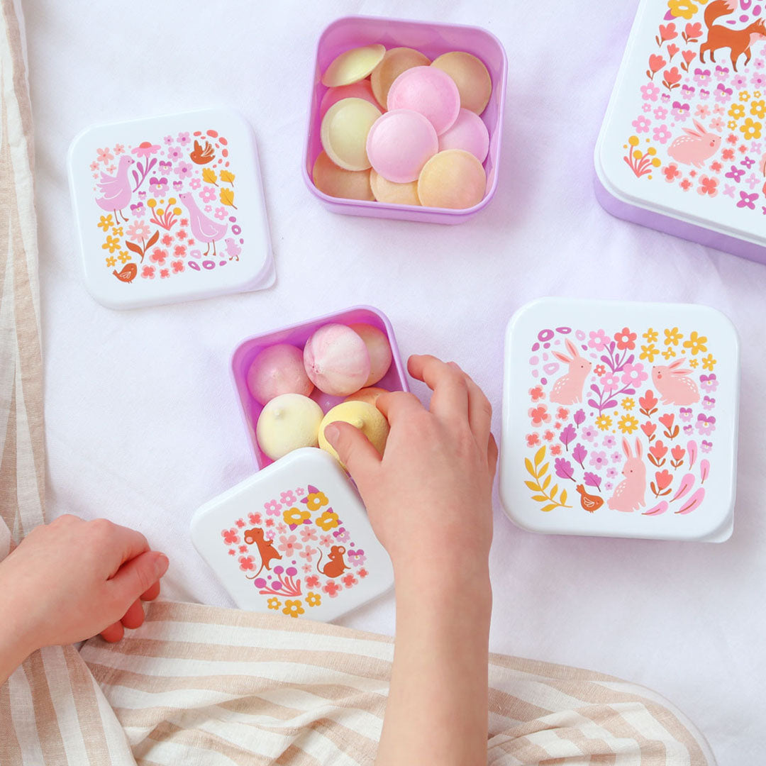 Een kind pakt snoepjes uit een kleine paarse A Little Lovely Company lunchbox met bloemen- en dierenpatroon, omringd door andere lunchboxen.