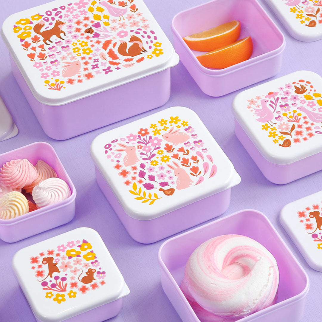 Meerdere paarse A Little Lovely Company lunchboxen met bloemen- en dierenpatroon, gevuld met diverse snacks op een paarse ondergrond.
