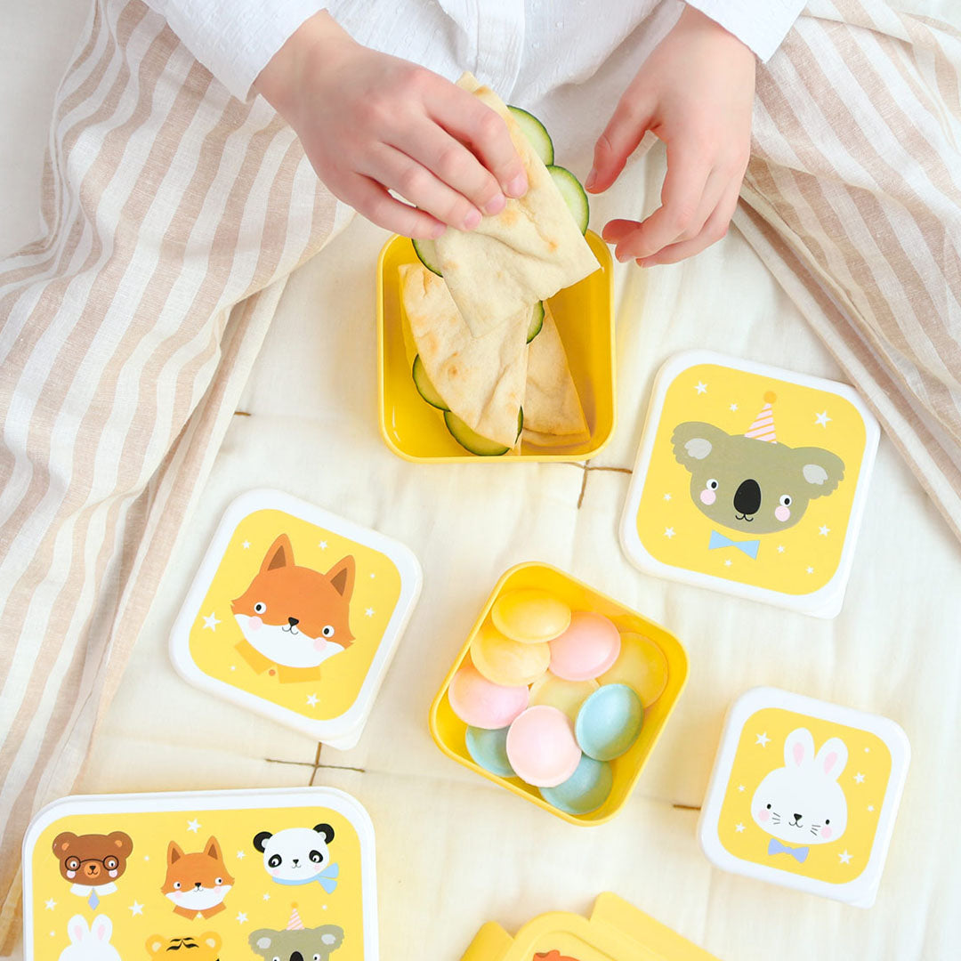 Een kind vult een gele lunchbox uit de A Little Lovely Company broodtrommelset met dierenvrienden en sterren met eten.