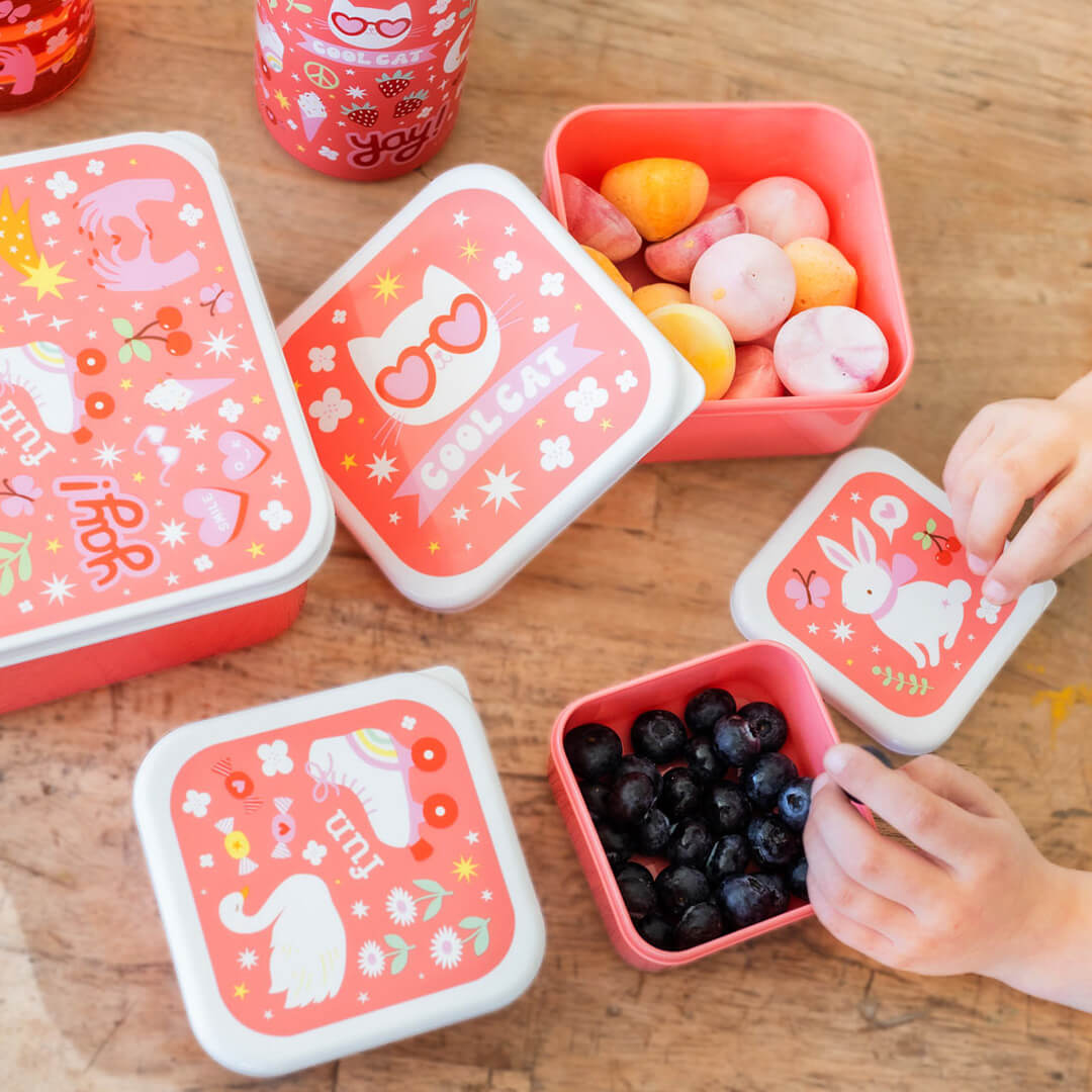 A Little Lovely Company lunchboxen op een houten tafel, gevuld met fruit en snoep, met kinderhanden die blauwe bessen pakken.