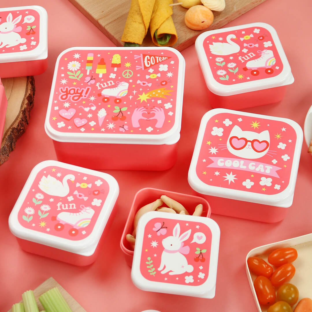Meerdere A Little Lovely Company lunchboxen op een roze ondergrond, gevuld met diverse snacks.