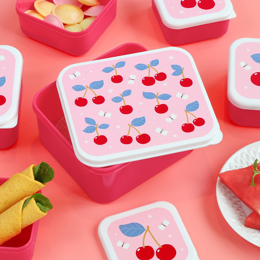 Een open roze A Little Lovely Company broodtrommel voor kinderen met kersenpatroon op de deksel, omringd door snacks op een roze ondergrond.