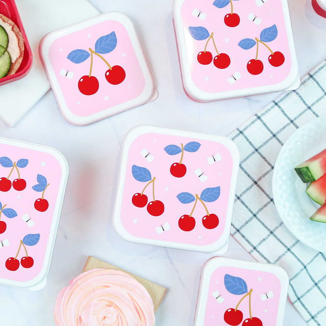Meerdere A Little Lovely Company broodtrommeldeksels voor kinderen met kersenpatroon, liggend op een lichte ondergrond met een theedoek en snacks.