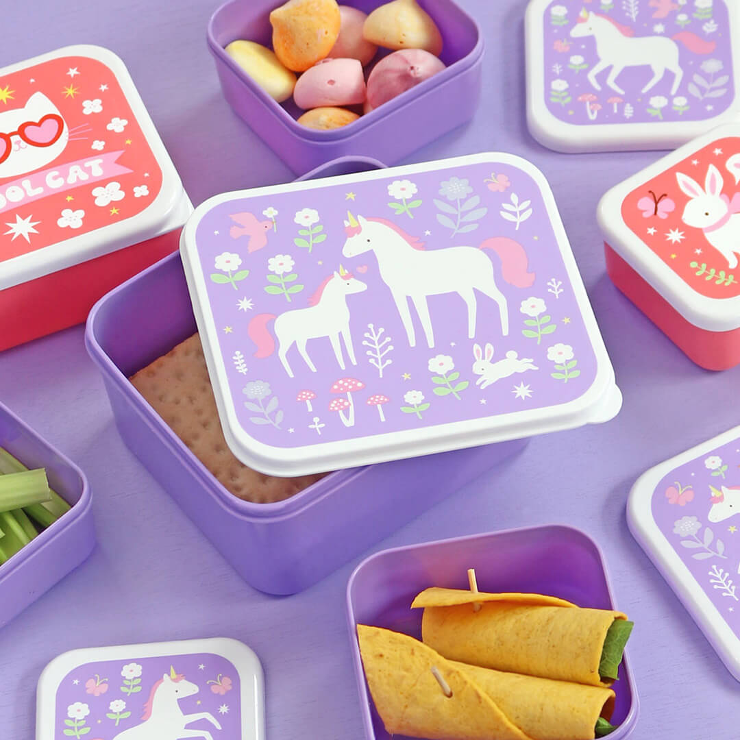 Een open paarse A Little Lovely Company lunchbox met een eenhoornpatroon, gevuld met een cracker, omringd door andere lunchboxen met snacks.