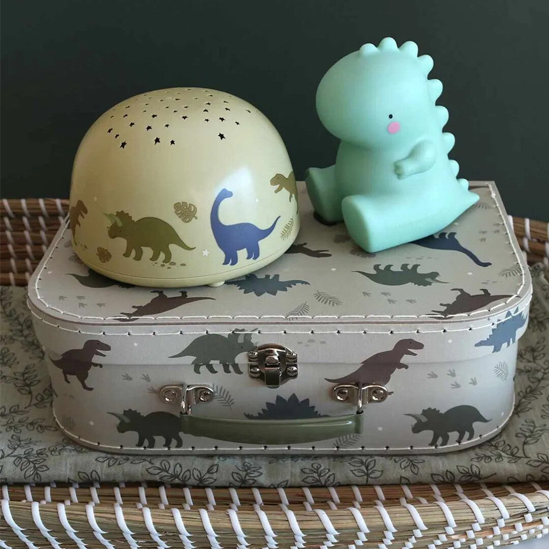 A little lovely company beige nachtlamp en sterprojector naast een groene dinosaurus nachtlamp op een koffer met dinosauruspatroon.
