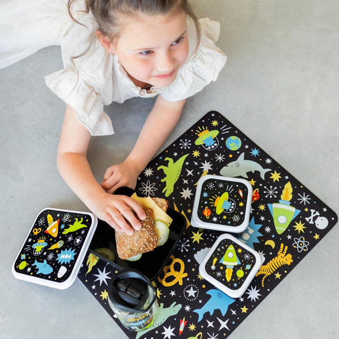 Een kind met een A Little Lovely Company placemat met galaxy patroon en bijpassende lunchboxen.