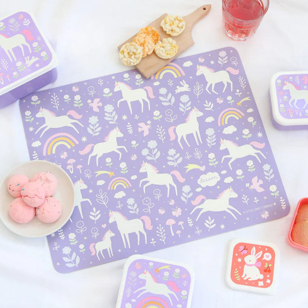 De paarse eenhoorn placemat van a little lovely company ligt op tafel met bijpassende lunchboxen en snacks.