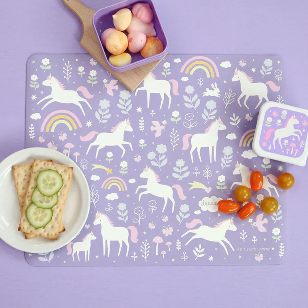 De paarse eenhoorn placemat van a little lovely company ligt op tafel met een bordje crackers en een bakje fruit.