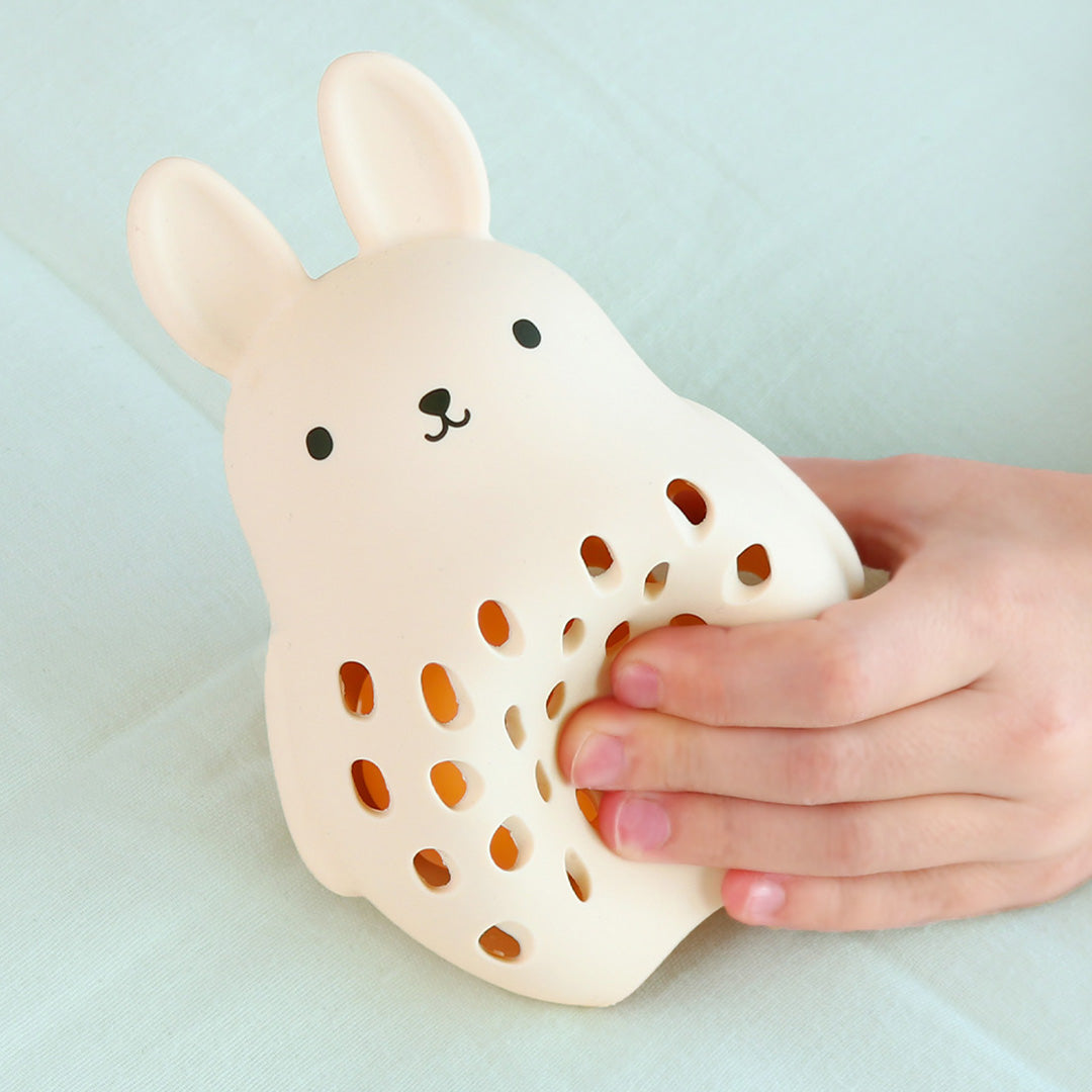 Kinderhanden houden een beige a little lovely company konijn rammelaar vast.