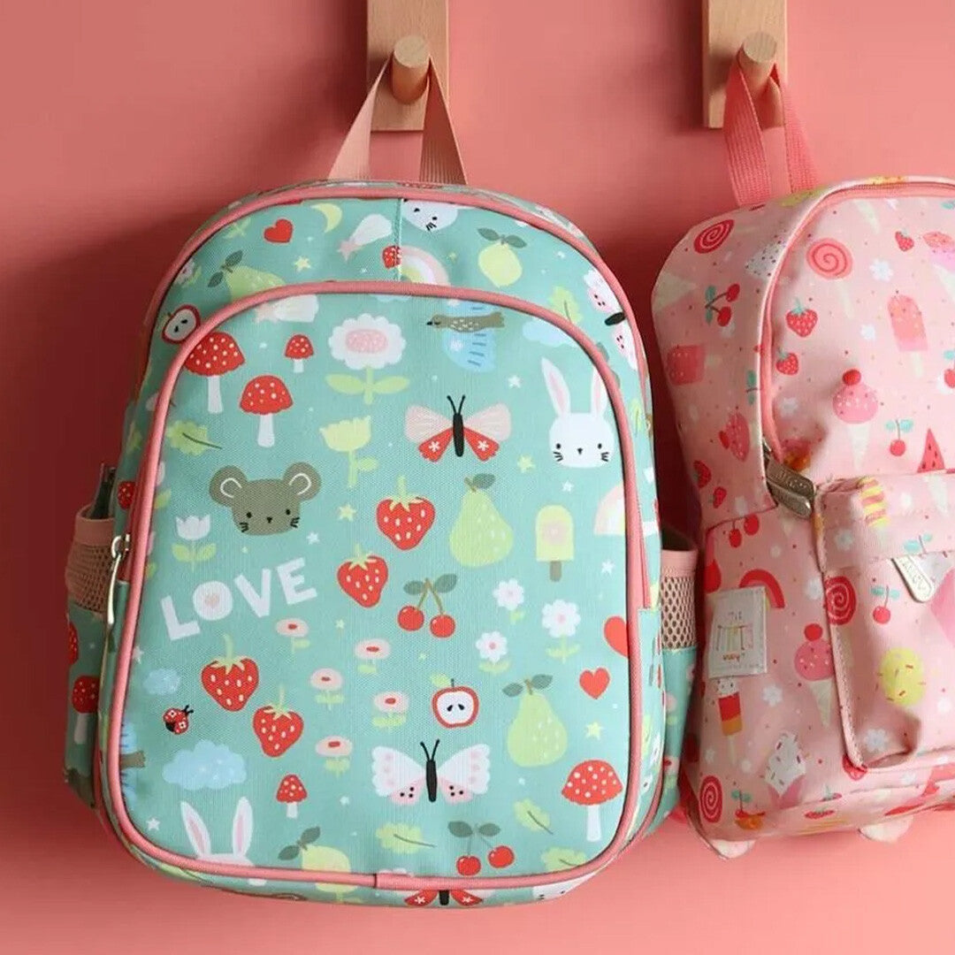 A Little Lovely Company groene kinderrugzak met dieren- en fruitpatroon hangt naast een roze rugzak aan een roze muur.