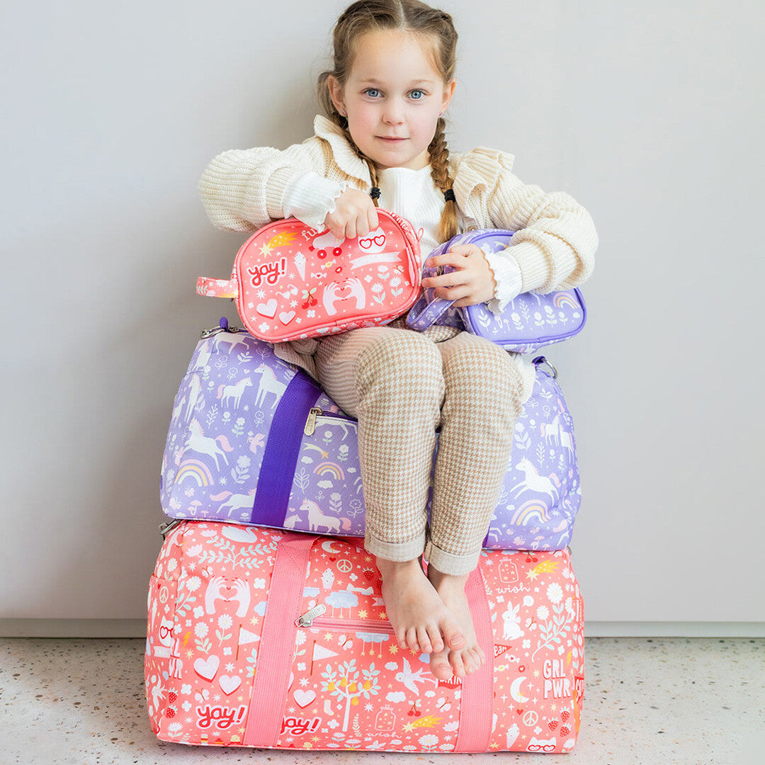 Meisje zit op een stapel a little lovely company bagage, met de roze toilettas en een paarse tas.