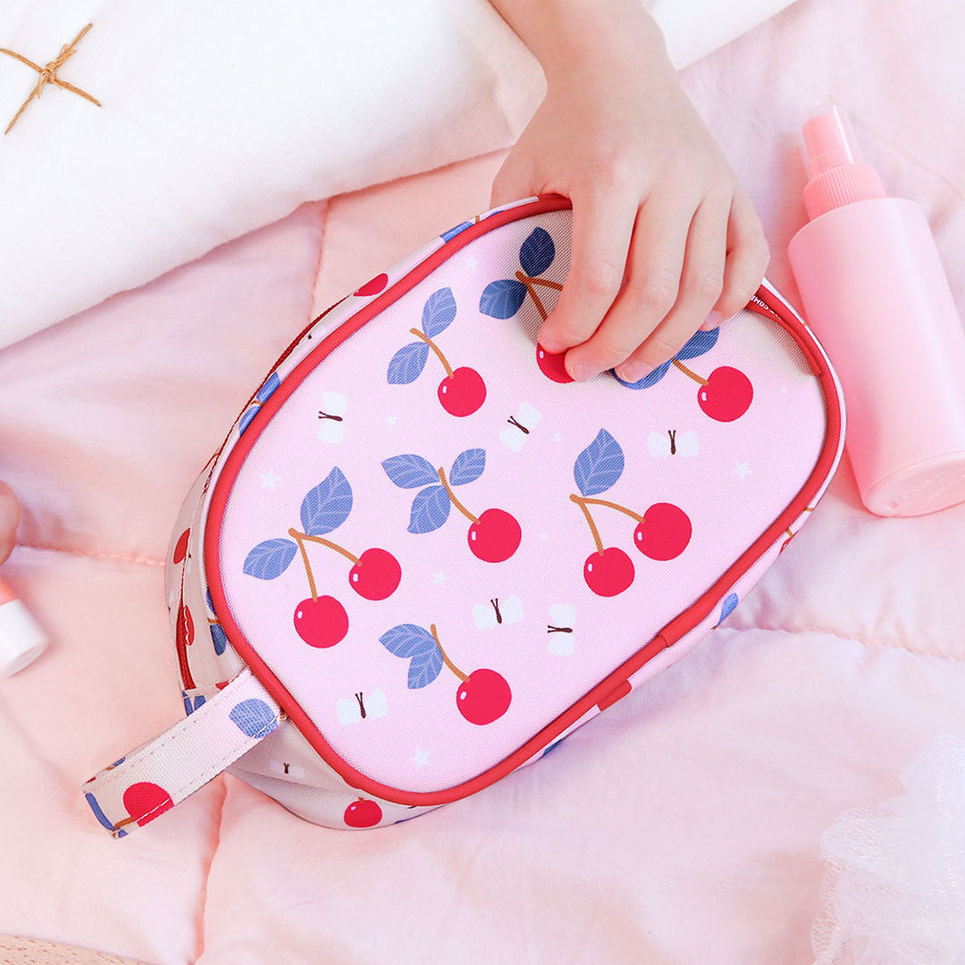 Een kinderhand houdt de a little lovely company roze toilettas met kersenpatroon vast op een roze deken.