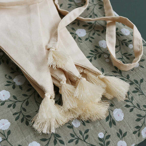 Detail van de crème stoffen vlaggenslinger met kwastjes, liggend op een groen doek met bloemenpatroon.