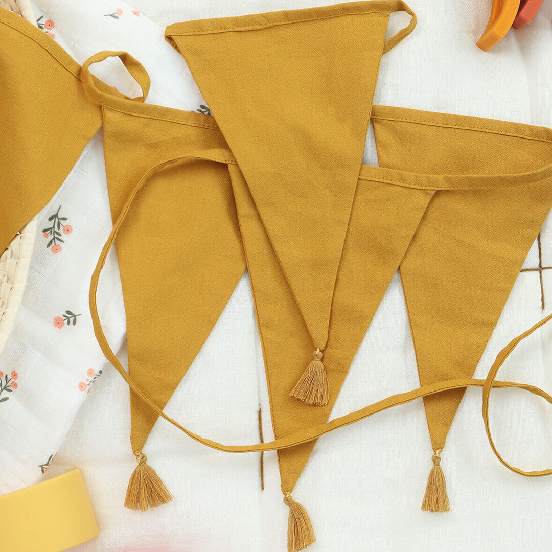 Detail van een a little lovely company okergele stoffen slinger met kwastjes, liggend op een witte ondergrond.