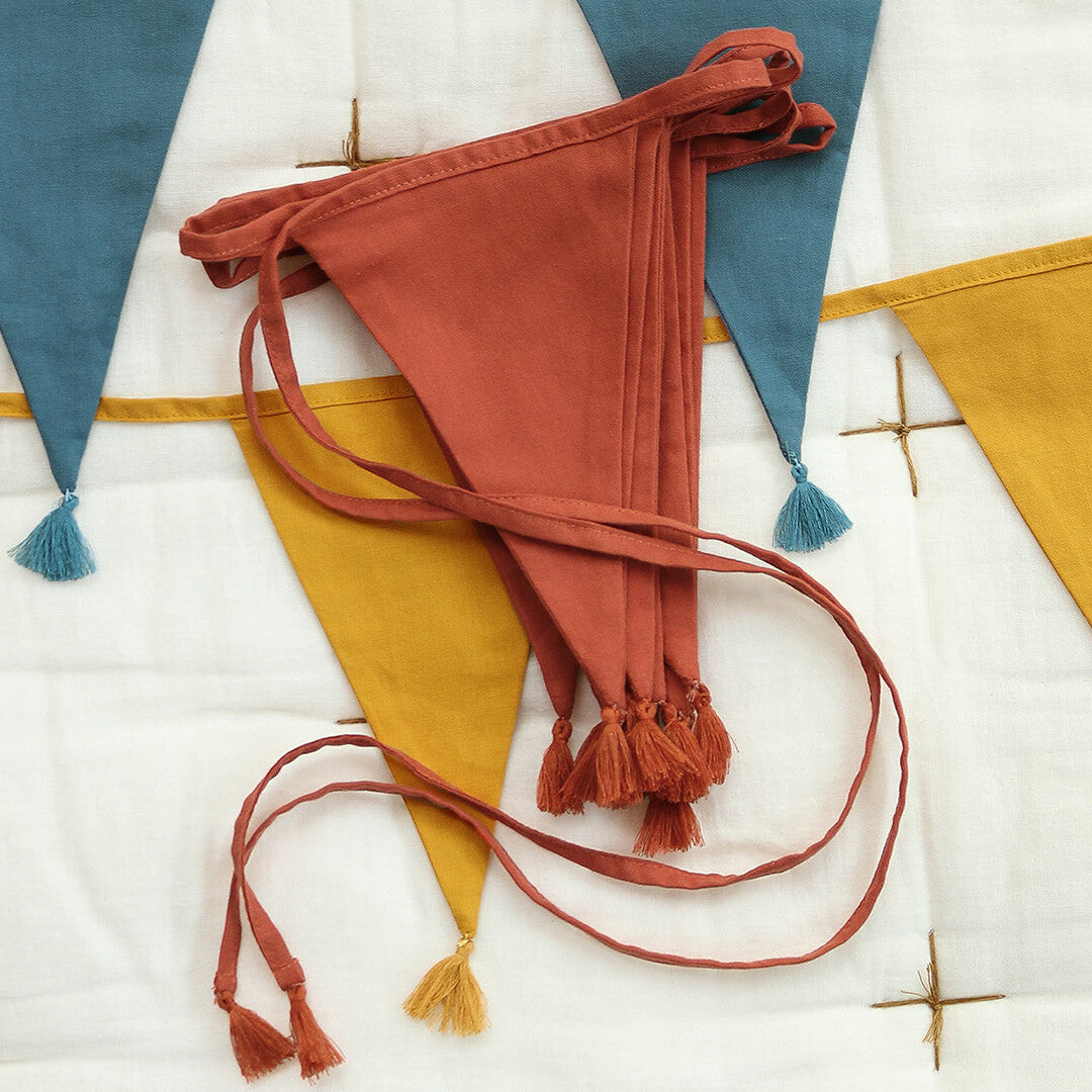 Een opgevouwen terracotta stoffen slinger met kwastjes, met delen van blauwe en okergele slingers op de achtergrond.
