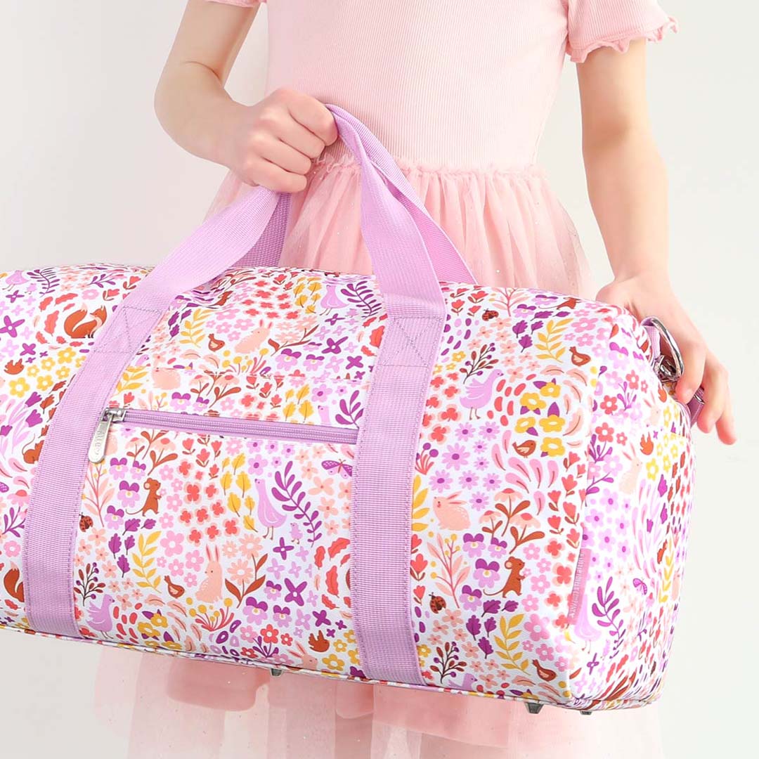 Een kind in een roze jurk houdt de a little lovely company kinder weekendtas met bloemen- en dierenprint vast.