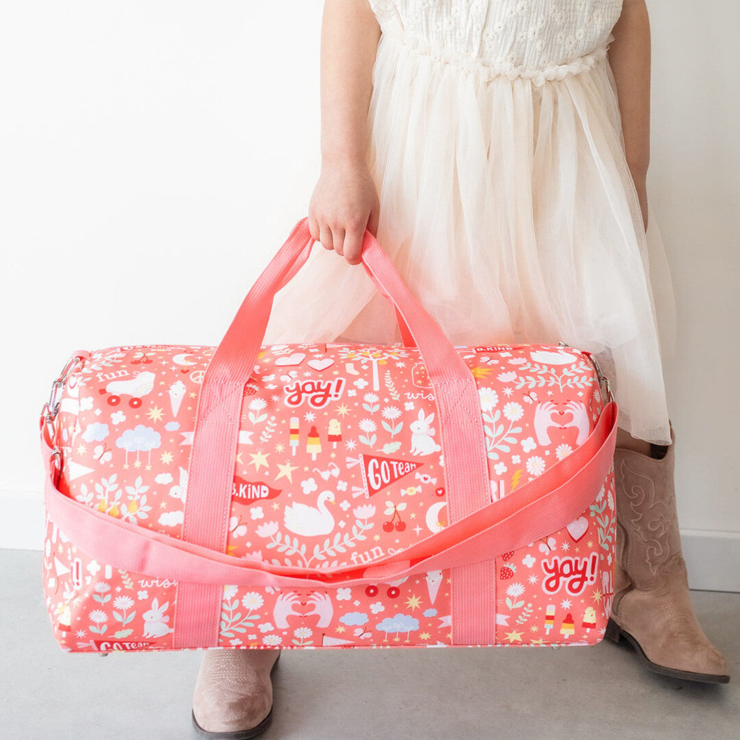 Een kind houdt de a little lovely company roze weekendtas met vrolijke print vast aan de handvatten.