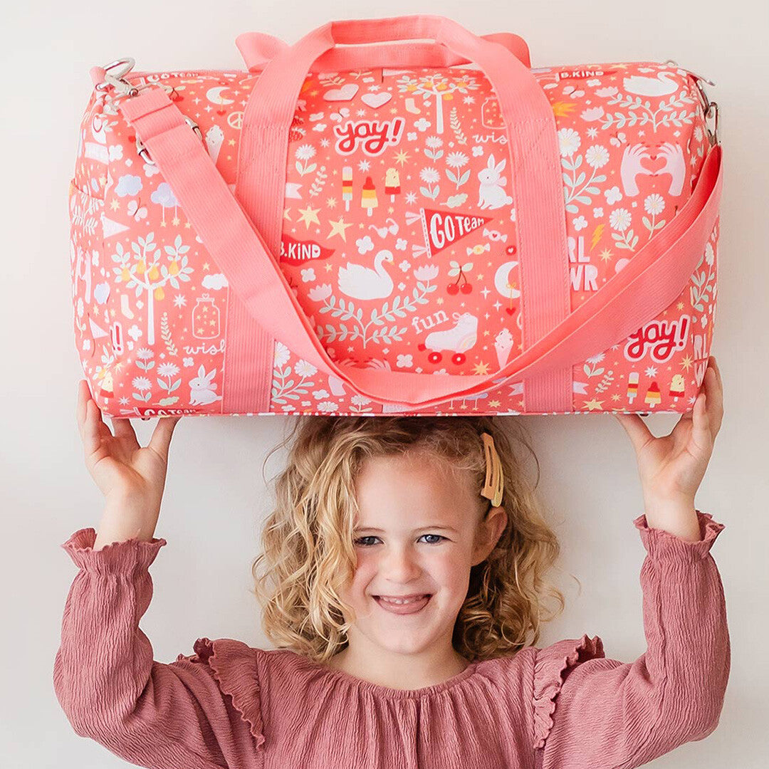 Een lachend kind balanceert de a little lovely company roze weekendtas met vrolijke print op haar hoofd.