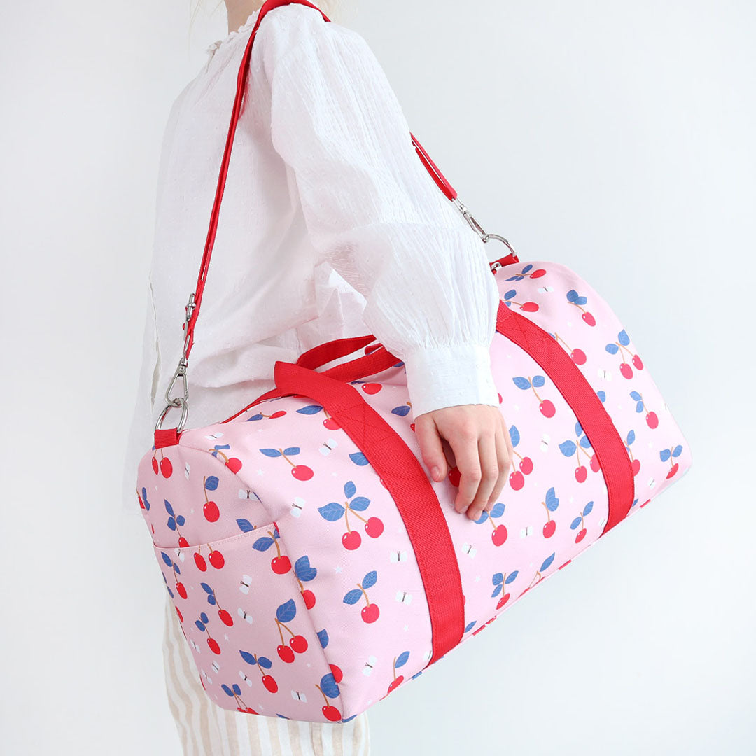 Een persoon draagt de roze a little lovely company kinder weekendtas met kersenprint over de schouder.