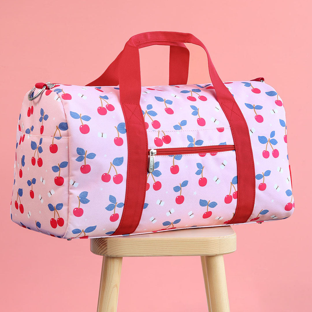 Een roze a little lovely company kinder weekendtas met kersenprint en rode hengsels staat op een houten krukje voor een roze achtergrond.