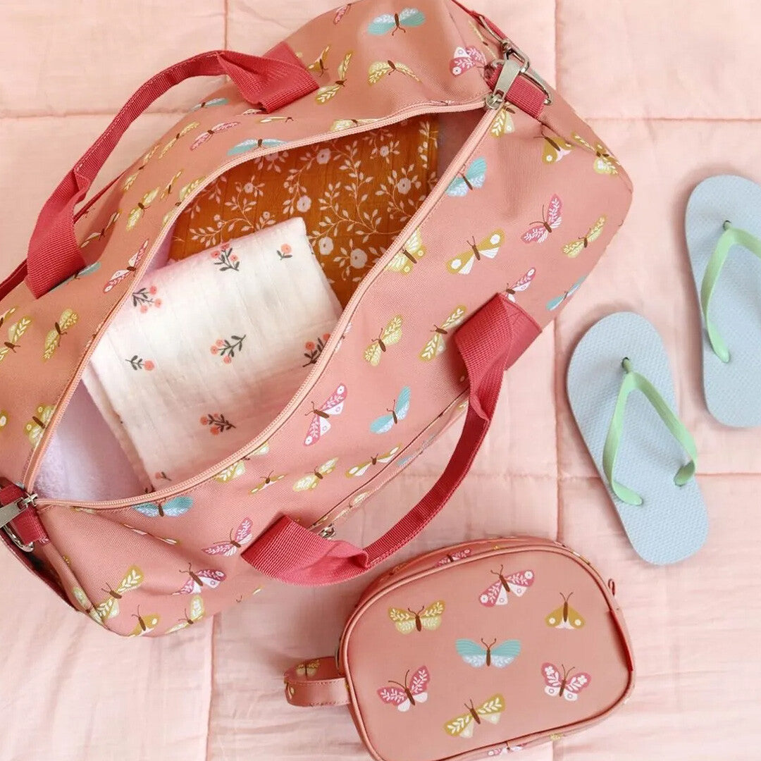 Een open a little lovely company roze kinderweekendtas met vlinderpatroon, gevuld met kleding, naast een bijpassend toilettasje en slippers.