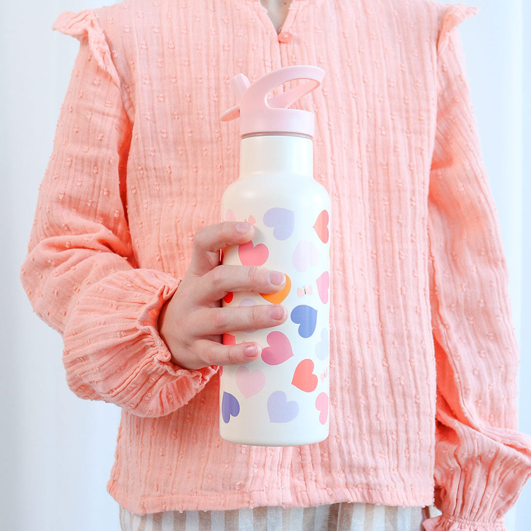 Een kind houdt de A little lovely company kinder drinkfles met hartjespatroon vast tegen een roze shirt.