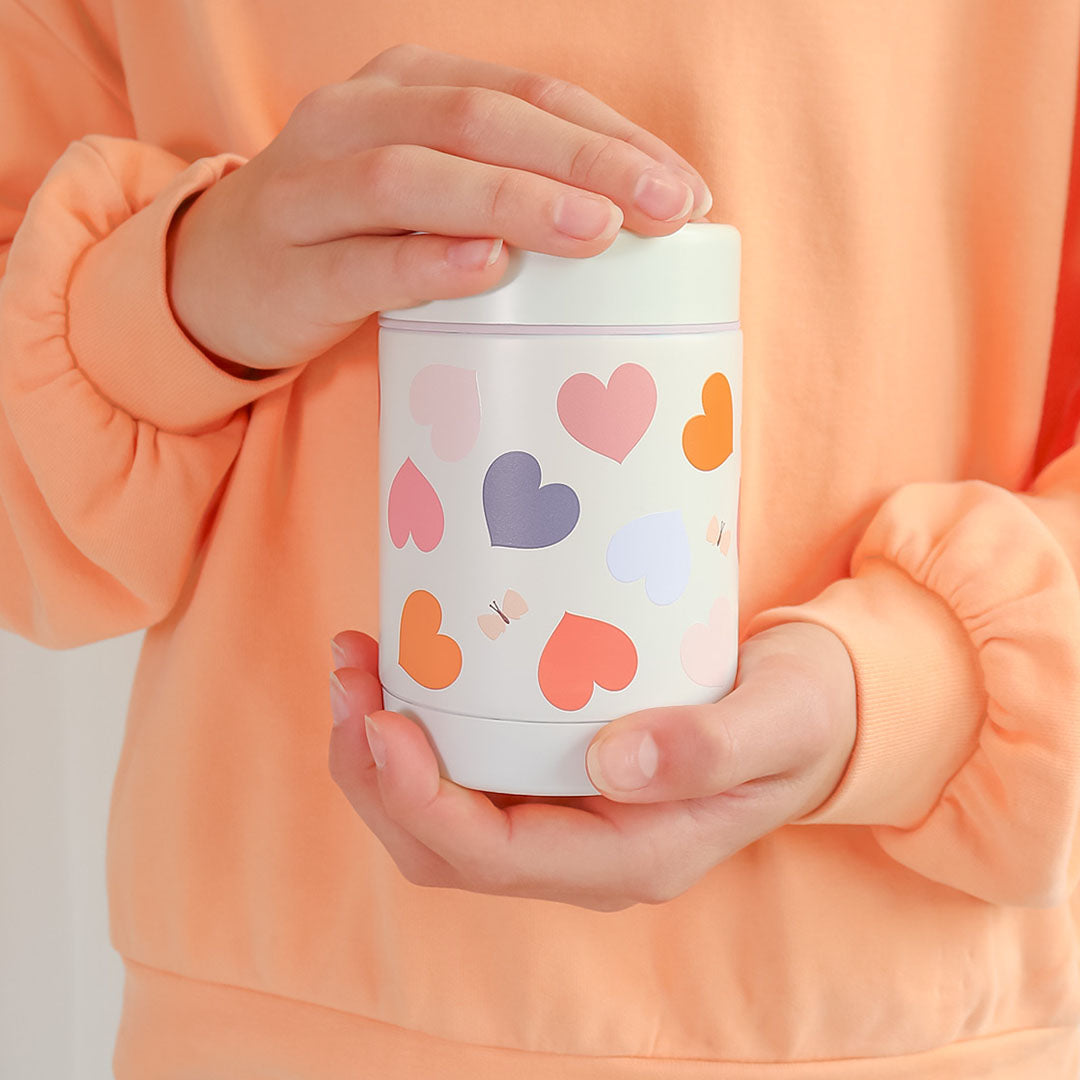 Twee handen houden de A Little Lovely Company RVS voedselpot thermos met hartjespatroon vast.