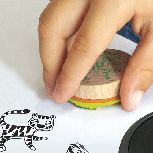 Een kinderhand drukt een Aladine houten tijgerstempel op wit papier, met gestempelde tijger en zebra ernaast.