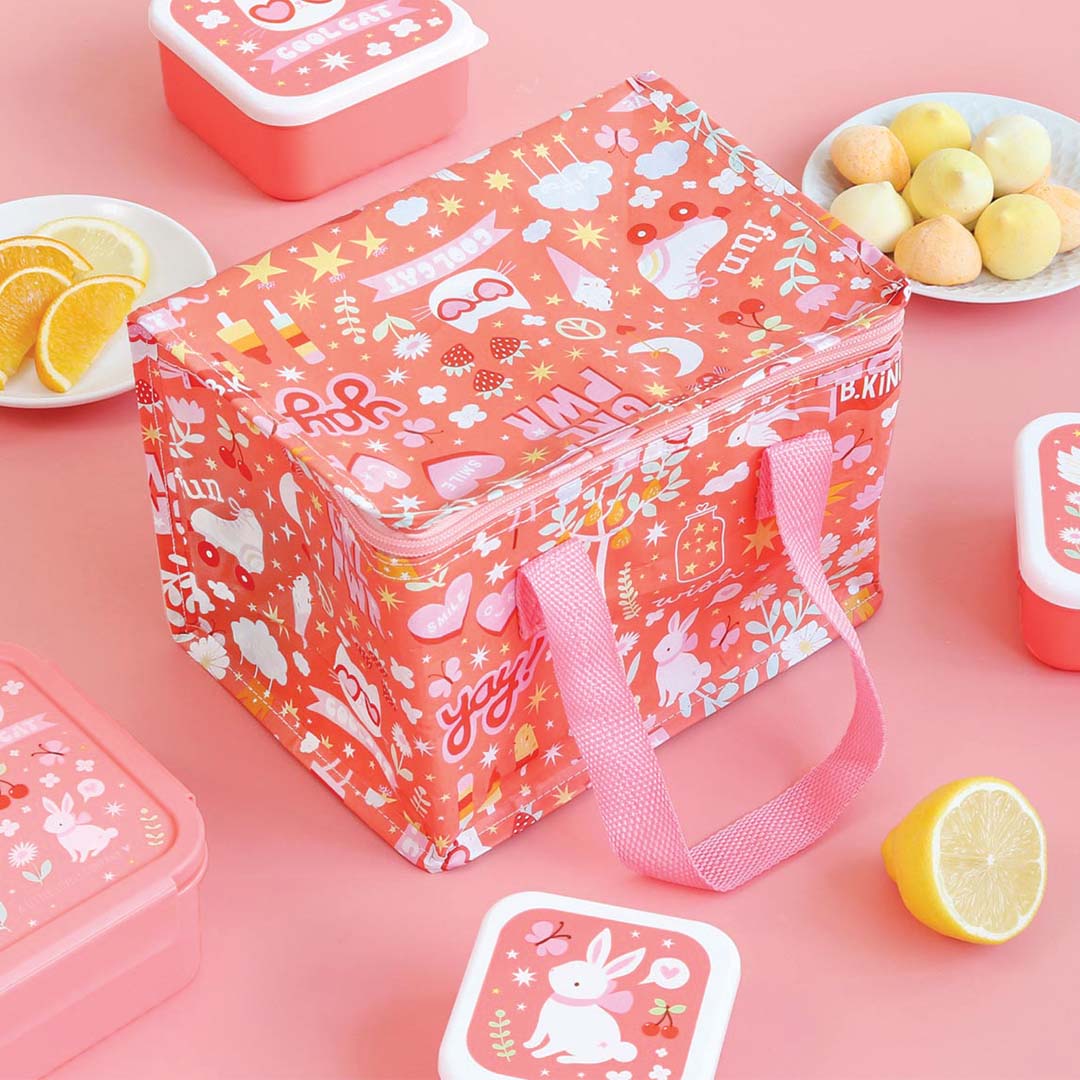 Een gesloten oranje a little lovely company koeltas met vrolijk patroon, omringd door kleine lunchboxen en snacks op een roze ondergrond.