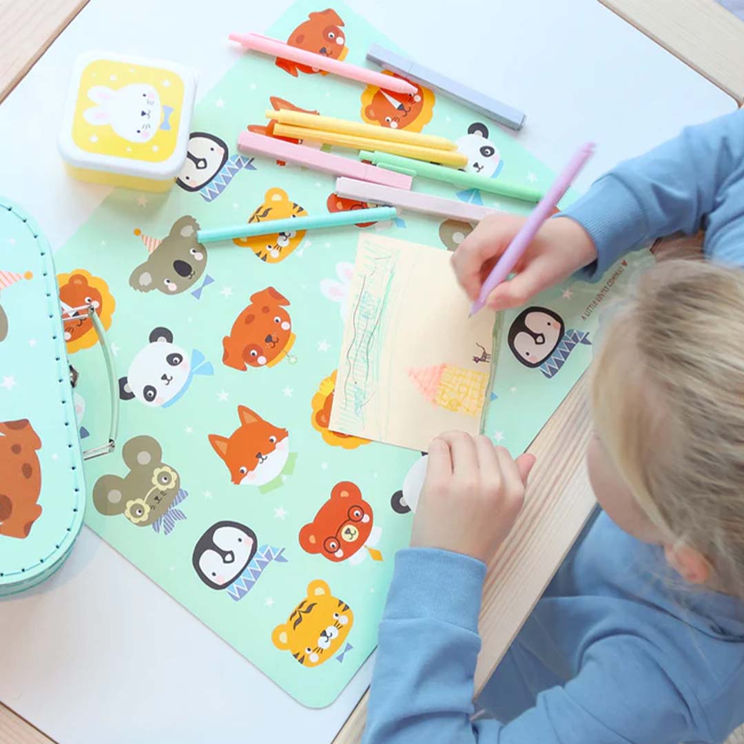 Kind tekent aan tafel op een A Little Lovely Company placemat met dierenvriendjes, omringd door kleurpotloden.