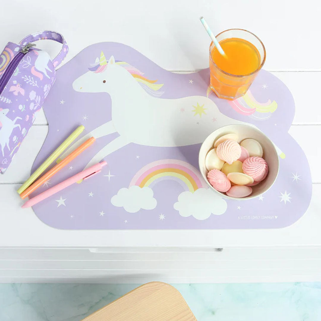 A Little Lovely Company placemat met eenhoorn en regenboog op een tafel met een glas sap en snoepjes.