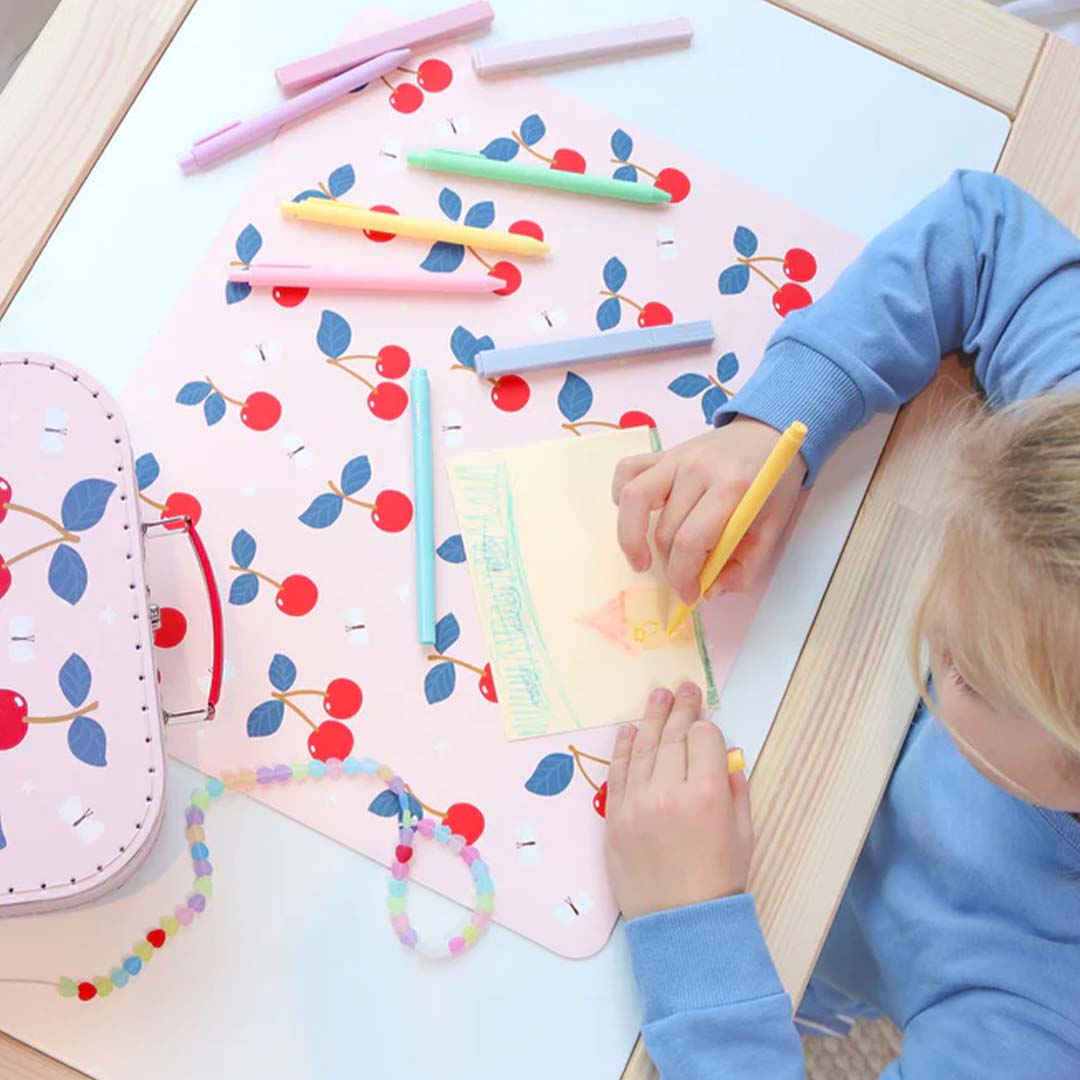 Een kind tekent op een A Little Lovely Company roze placemat met kersenpatroon, omringd door pennen en een koffer.