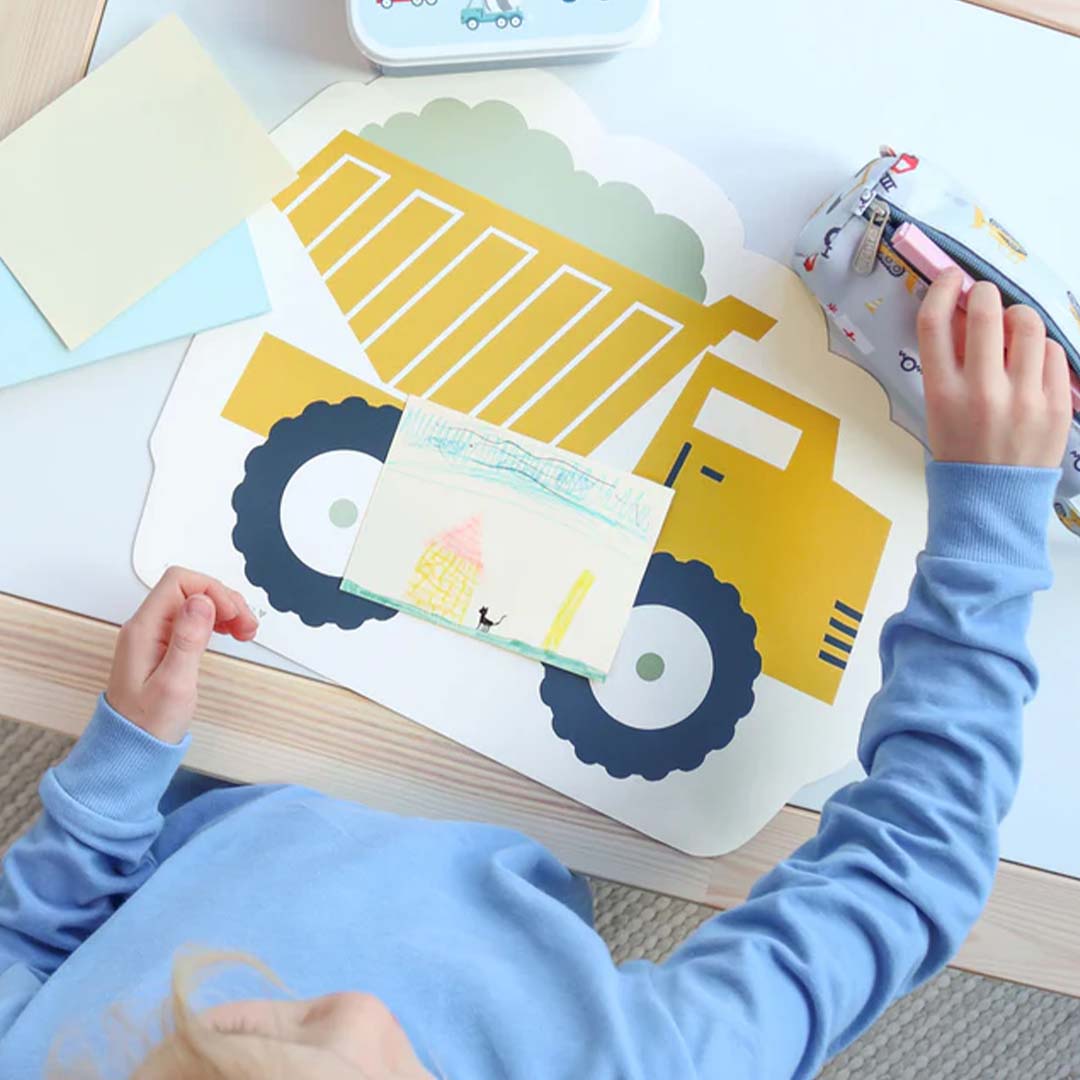 Een kind tekent aan tafel op een gele kiepwagen placemat van A Little Lovely Company.