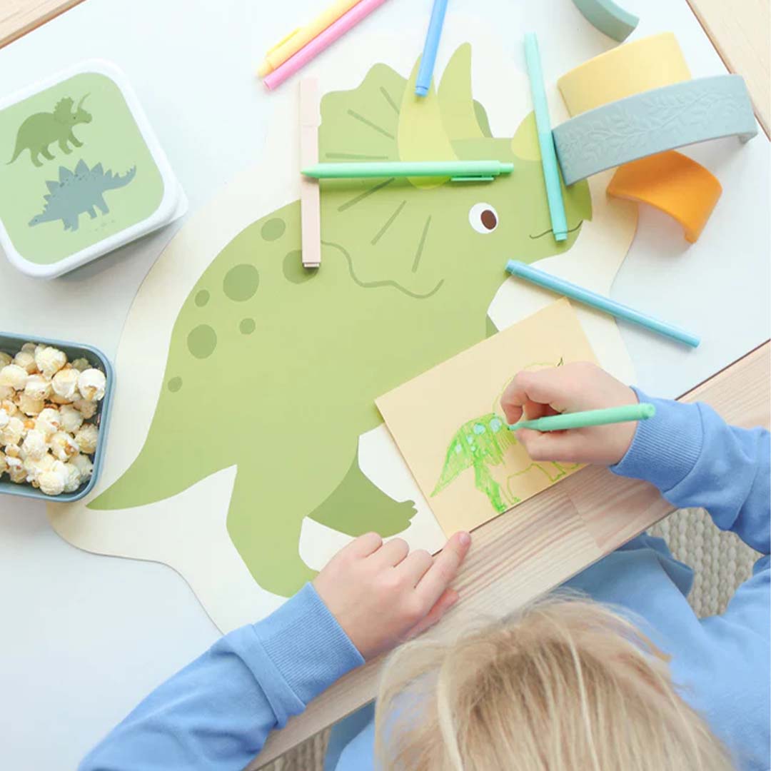 Een kind tekent aan tafel naast de A Little Lovely Company placemat in de vorm van een groene dinosaurus.