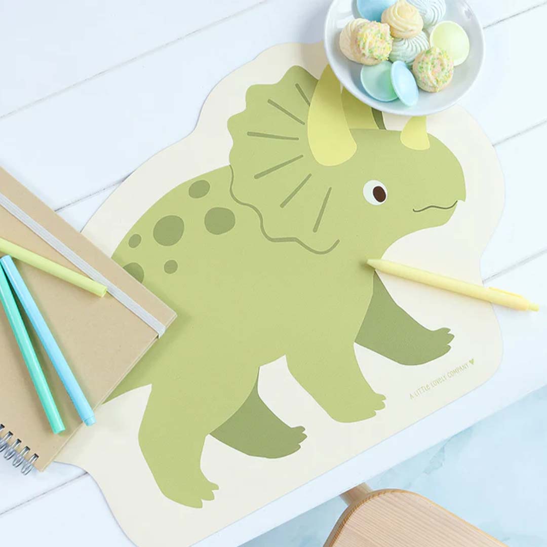 De A Little Lovely Company placemat in de vorm van een groene dinosaurus op een witte tafel met schrijfwaren en snoepjes.