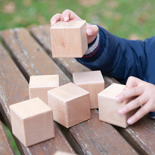 Een kinderhand houdt een Andreu Toys houten sensorisch blok vast, met andere blokken op een houten bank.
